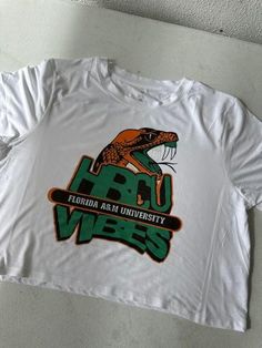 Choose from a variety of FAMU-inspired designs: Available Sizes and Colors:  Adult Small, Medium, Large, XL, XXL Black, White, Orange & Green   Don't see a design or color you're looking for? Feel free to message us for customs! We're open to all ideas! Fit: Unisex fit, true to size Care Instructions: Machine wash cold, inside out Tumble dry low Do not iron directly on the design Inspiration: "Each design in our FAMU collection is crafted with love for the Rattler community. Whether you're celebrating homecoming, showing school pride, or gifting to a fellow alum, these shirts are the perfect representation of Florida A&M's legacy." White Screen Print Top For Fan Conventions, Funny Tops With Sublimation Print For Fans, Funny Fan Merchandise Tops With Sublimation Print, Fun Graphic Print Tops For Fan Conventions, Trendy Custom Print Tops For Fan Merchandise, Letter Print Short Sleeve Tops For Fan Conventions, Short Sleeve Tops With Letter Print For Fan Conventions, All Ideas, We're Open