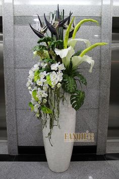 a white vase filled with lots of flowers on top of a floor next to a wall