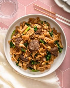 a white bowl filled with noodles and meat next to chopsticks on a pink table