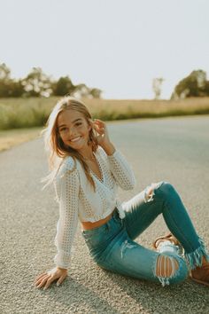 a beautiful young woman sitting on the side of a road wearing jeans and a white top