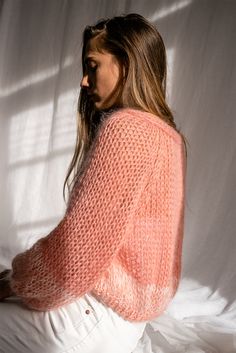 a woman sitting on top of a bed wearing a pink knitted sweater and white pants