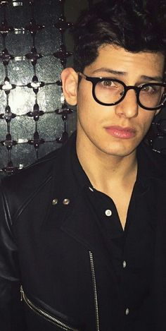 a young man wearing glasses standing in front of a wall with metal squares on it