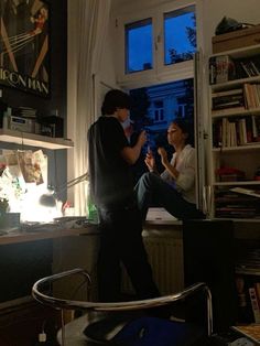 two people standing in front of a window with bookshelves and shelves behind them