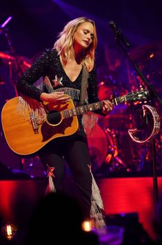 a woman holding a guitar while standing in front of a microphone and singing on stage