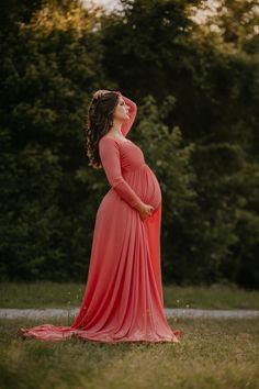 a pregnant woman in a red dress is standing on the grass with trees behind her