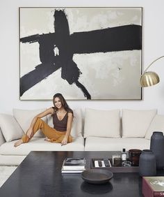 a woman sitting on top of a couch in front of a large abstract painting over a coffee table