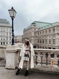 a woman wearing a white coat and hat standing on a bridge in front of some buildings