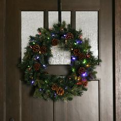 a wreath with lights and pine cones hanging from the front door on a wooden door