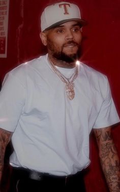 a man with tattoos and a white t - shirt standing in front of a red wall