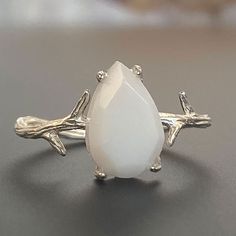 "For fans of minimalist nature-inspired jewelry, we created our stand-out Mother Nature Silver breastmilk ring. It pairs together a branch-like sterling silver ring band with an exquisite yet small pear-shaped faceted breastmilk stone. Go with your natural breast milk or combine your breast milk with opal effect flakes or pearl powder. Opalizing your breast milk adds a pale, milky opal appearance to your breastmilk stone that catches the light and reflects pastel rainbow hues, while adding pearl Minimalist White Moonstone Ring In Sterling Silver, Delicate White Moonstone Ring In Sterling Silver, Unique Adjustable White Moonstone Ring, Unique White Adjustable Moonstone Ring, Delicate White Hypoallergenic Ring, Delicate Hypoallergenic White Rings, Delicate Handmade White Rings, White Teardrop Sterling Silver Rings, Delicate White Moonstone Gemstone Ring