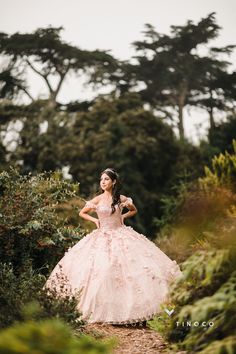 a woman in a pink dress standing on a dirt path surrounded by bushes and trees