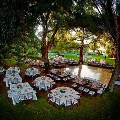 tables and chairs are set up for an outdoor dinner under the shade of trees near a body of water