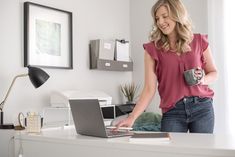 a woman is holding a coffee mug and looking at her laptop while standing in front of a desk