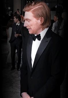 a man in a tuxedo and bow tie standing next to other people at an event