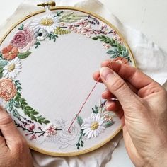 two hands are stitching a flowered embroidery on a piece of white fabric with a needle