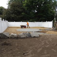 a man with a wheelbarrow in the middle of a graveled area next to a white fence