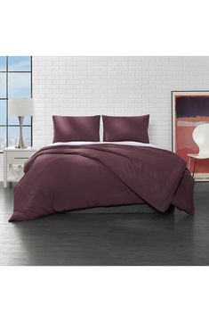 a bed with purple sheets and pillows in a white brick wall room next to a painting