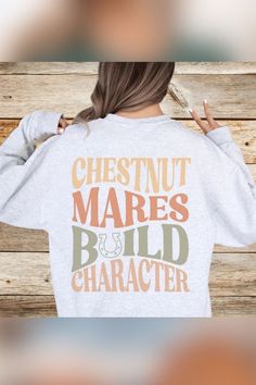 A woman with long brown hair faces away, wearing a light gray sweatshirt that reads "CHESTNUT MARES BUILD CHARACTER" in large, earthy tones. She holds up one arm, making a peace sign with her hand, standing in front of a rustic wooden wall. Chestnut Mare, Cowgirl Outfits