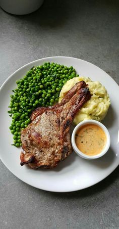 a white plate topped with meat, mashed potatoes and green peas next to a small bowl of sauce