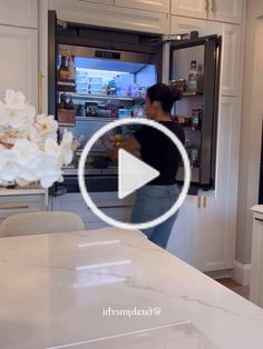 a woman standing in front of a refrigerator with flowers on the counter and an open freezer door