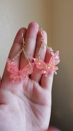 a hand holding two pink butterfly shaped earrings in it's left hand, with chain attached to them