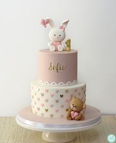 a pink and white two tiered cake with a teddy bear sitting on the top