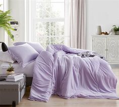 a bed covered in purple sheets and pillows next to a potted plant on top of a dresser