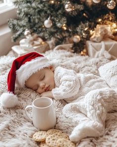 a baby wearing a santa hat sleeping on a blanket next to a christmas tree with cookies