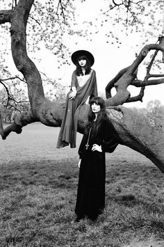 two women standing in front of a tree with their arms around each other, both wearing long dresses and hats