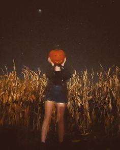 a woman standing in the middle of a corn field holding an orange pumpkin up to her face