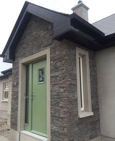 a green door is on the side of a stone building with two windows and a black roof