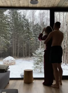 two people standing in front of a window looking out at the snow covered forest outside