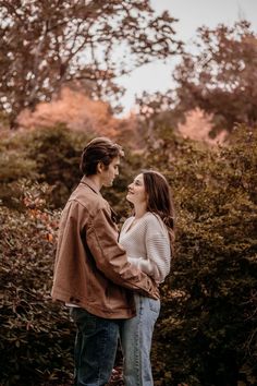 two people standing next to each other in front of some bushes and trees with one person holding the other's hand