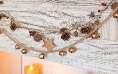 a string of pine cones and bells hangs on a white wooden wall with a candle