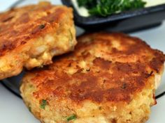 three crab cakes are sitting on a plate with some greens in the bowl behind them