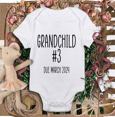 a white onesuit with the words grandchild 3 due march 2021 on it and a teddy bear next to it