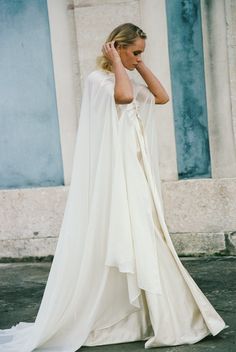 a woman in a white wedding dress with a cape on her head and hands behind her head