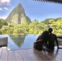 two people are sitting on the edge of a pool looking out at mountains and trees