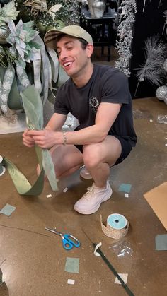 a man kneeling down next to a plant with scissors and tape on the ground in front of him