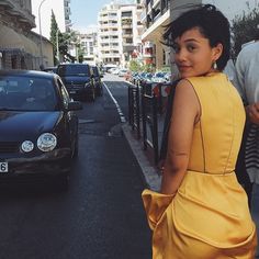 a woman in a yellow dress is walking down the street with cars and people behind her