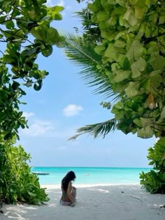 a woman sitting on the beach looking out at the blue water and green trees in front of her