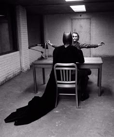 two people sitting at a table in an empty room with one person dressed as batman