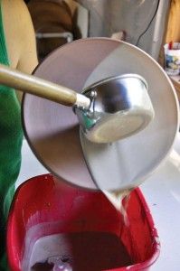 a person pouring liquid into a red bucket with a wooden spoon in it on a table