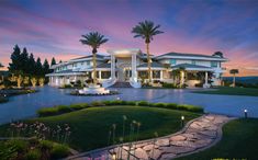 a large white house with palm trees in the front yard at dusk, surrounded by greenery and landscaping