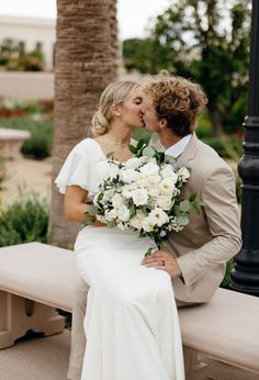 a bride and groom kissing while sitting on a bench in front of some palm trees