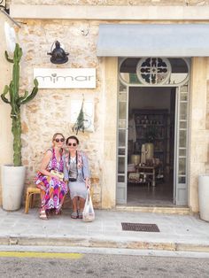 two women sitting on a bench in front of a building with a cactus and potted plant