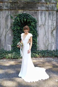 a woman standing in front of a wooden fence wearing a white dress and holding a bouquet