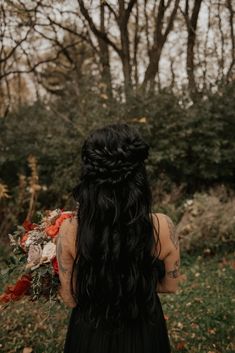 a woman with long black hair standing in front of trees and leaves, looking at the ground