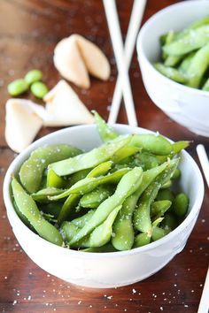 two white bowls filled with green beans next to chopsticks on a wooden table