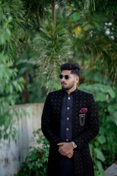 a man standing in front of some trees wearing a black suit and sunglasses with a flower on his lapel